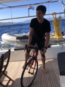 A smiling cyclist is riding a road bike on an ongoing catamaran sailing boat